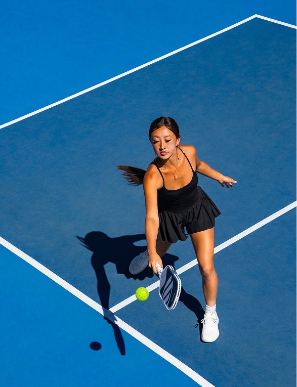 Bird's-eye view of a female pickleball player wearing an all-black athletic outfit preparing to strike a ball with a Apollo Axis pickleball paddle on a bright blue court.