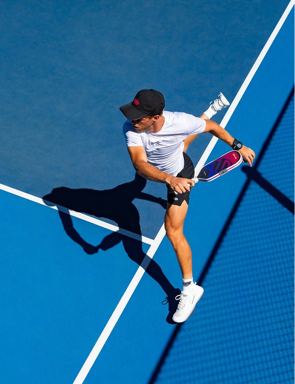 Overhead view of a male athlete running across a blue court, holding a gradient pink and purple Apollo Axis pickleball paddle.