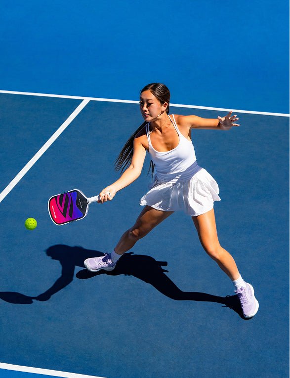 Action shot of a female pickleball player in a white athletic dress hitting a low forehand shot with a pink and purple gradient Apollo Axis pickleball paddle.