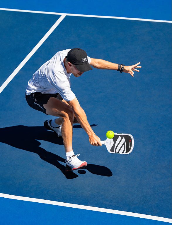 Overhead perspective of a male pickleball player bending low to return a yellow ball near the kitchen line with a black and white patterned Apollo Axis pickleball paddle.